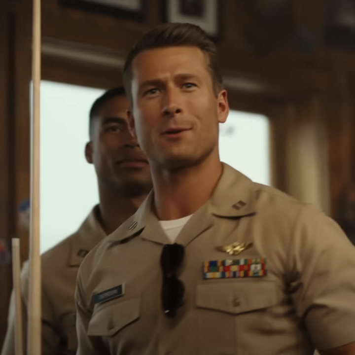 Glen Powell and Danny Ramirez in beige military uniforms, standing indoors with framed photos in the background