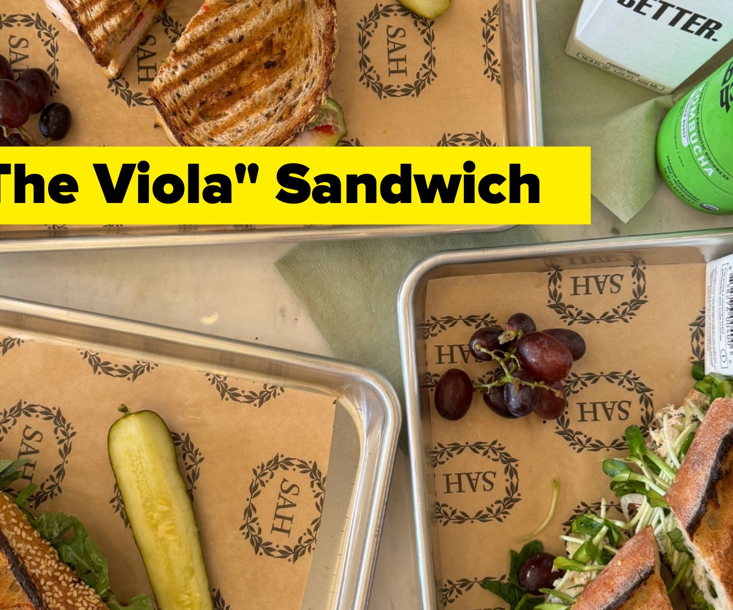 Three trays of deli sandwiches with pickles and grapes alongside boxed water and a bottled green drink on a table