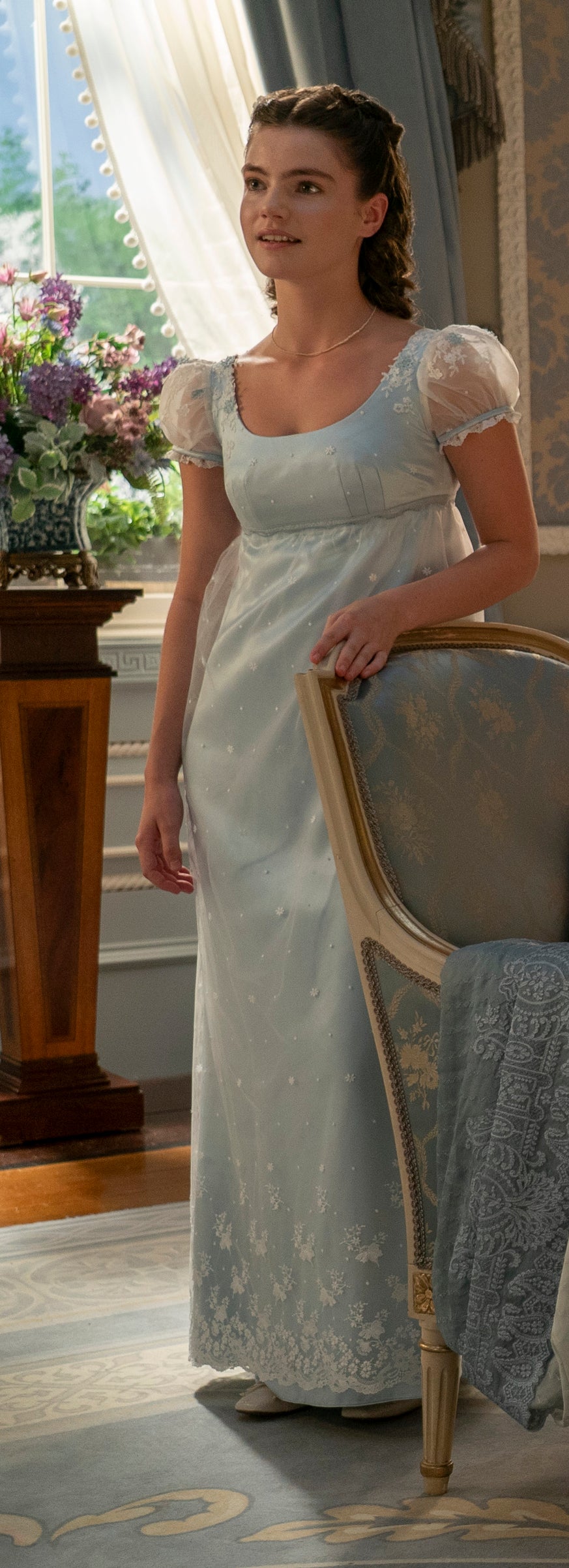 Hyacinth in an empire waist gown with sheer cap sleeves, leaning on a chair