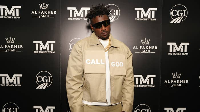Antonio Brown in a beige jacket with &quot;Call God&quot; printed on the front stands in front of a branded backdrop at an event