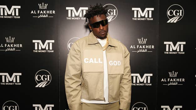 Antonio Brown in a beige jacket with "Call God" printed on the front stands in front of a branded backdrop at an event