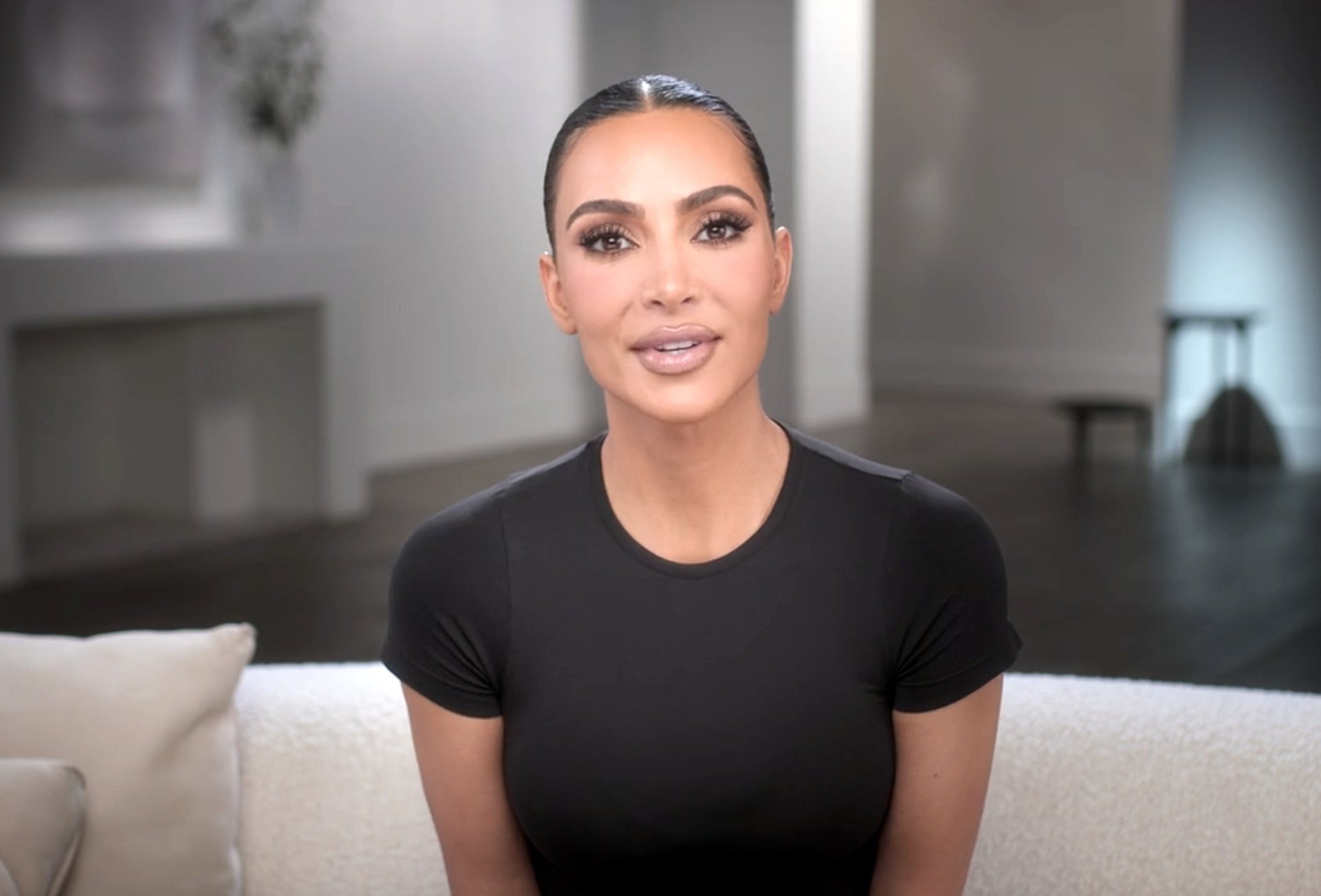 Kim Kardashian sits on a couch wearing a fitted top with her hair slicked back