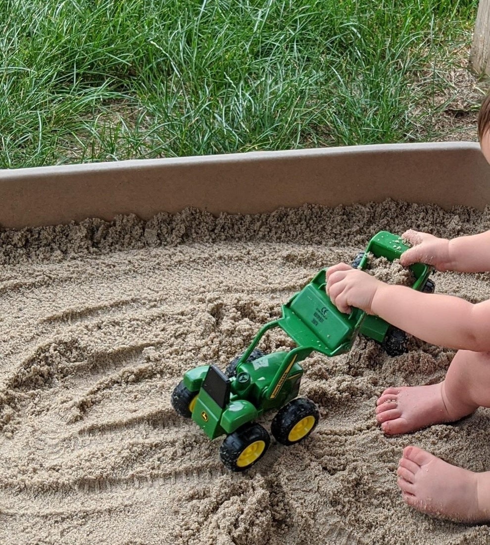 A child's hands and feet shown playing with a toy tractor in a sandbox
