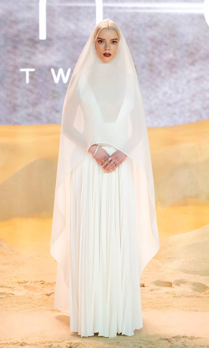 Anya Taylor-Joy on a red carpet wearing a modest, flowing white gown and a sheer white veil covering her head