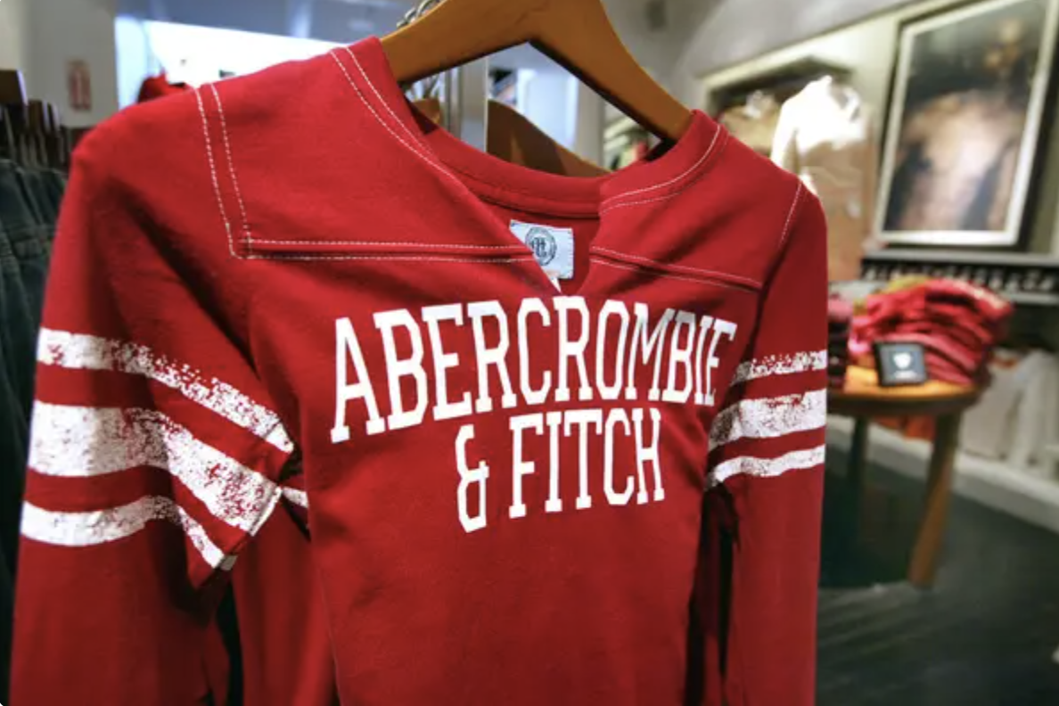 Red Abercrombie & Fitch sweatshirt with white stripes on sleeves, hanging on a wooden hanger in a store. Stack of shirts and framed pictures in the background