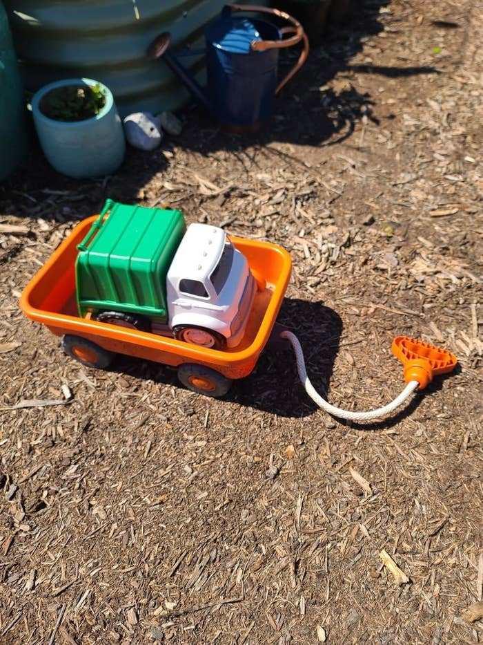 A small toy garbage truck with a green back in an orange trailer is outdoors on a dirt surface, with a gardening hose and plants nearby