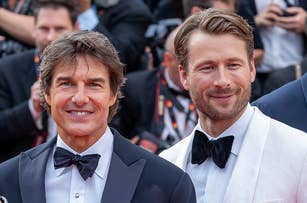 Tom Cruise and Glen Powell smile on the red carpet wearing tuxedos with bow ties. Photographers are visible in the background