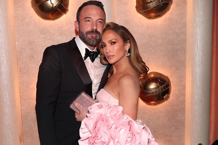 Ben Affleck and Jennifer Lopez pose together in formal attire, with Jennifer wearing a dramatic, floral-themed dress and holding a sparkling clutch