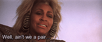 Tina Turner, wearing large spiral earrings and a futuristic outfit, smiles with the text "Well, ain't we a pair…" displayed at the bottom of the image