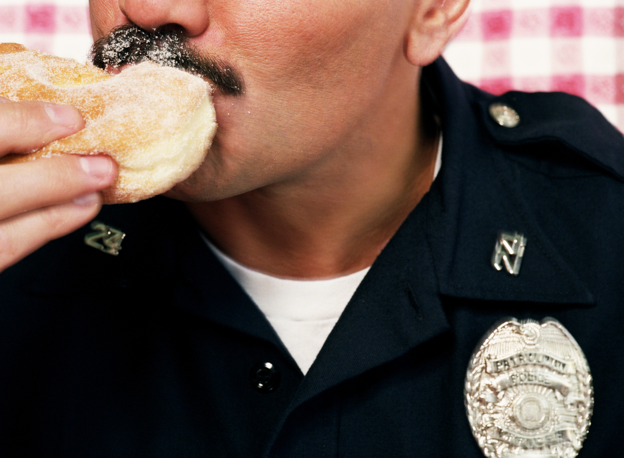 A person in a police uniform is eating a powdered donut. The face is partially obscured, showing a moustache