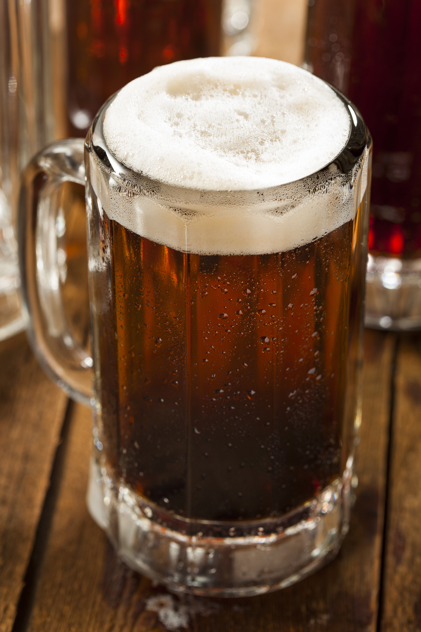 A frothy glass mug filled with dark beer on a wooden table, with other similar mugs visible in the background