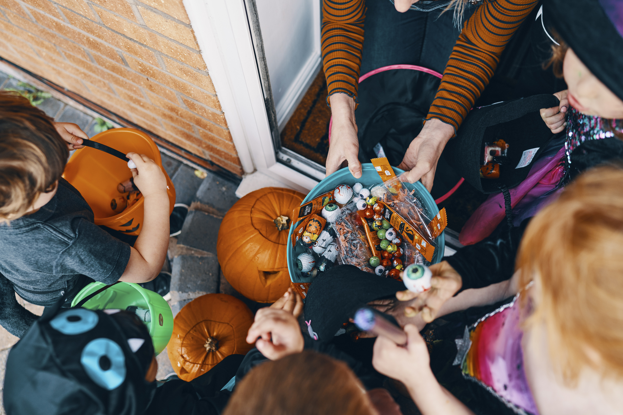 Children in costumes gather around a house entrance for trick-or-treating on Halloween, reaching into a bowl of assorted candies held by an adult