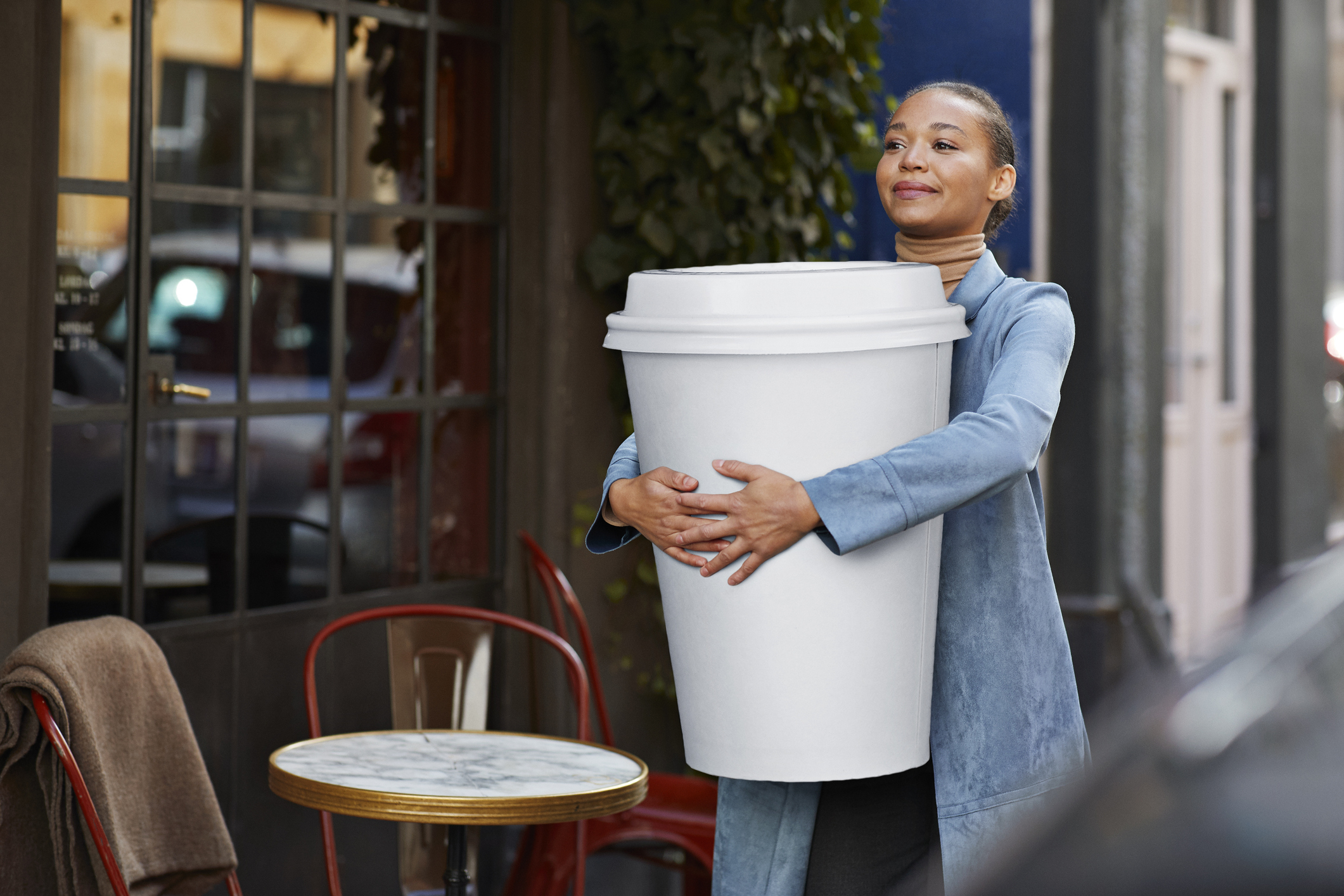 A woman stands outside, happily holding an oversized coffee cup that is much larger than a typical cup
