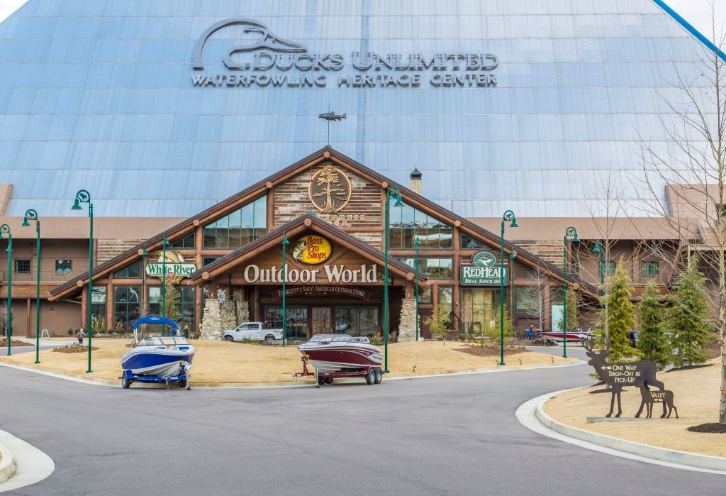 The image shows the entrance of a Bass Pro Shops Outdoor World store with boats parked in front. A large Ducks Unlimited Waterfowling Heritage Center sign is on the building's facade