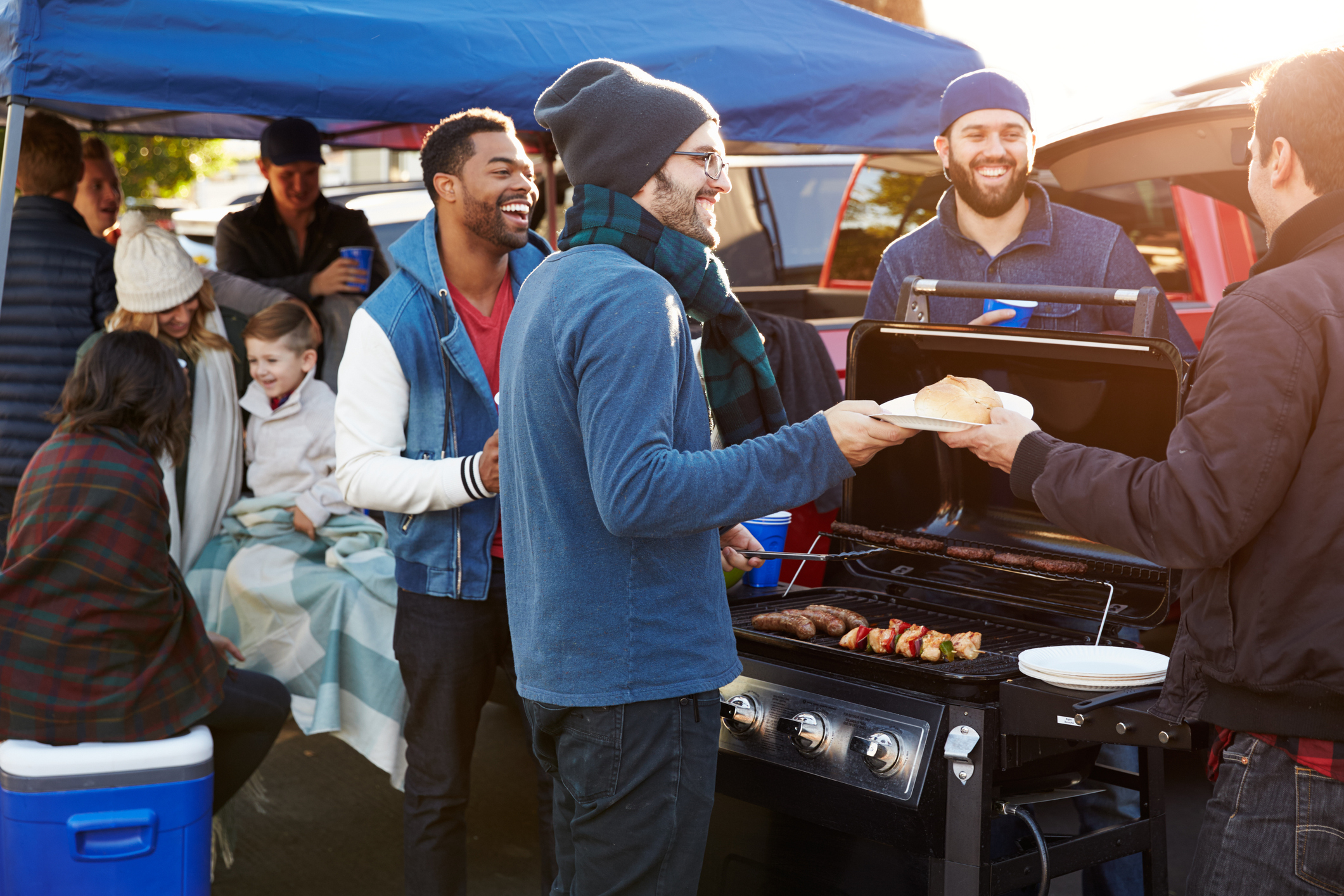 People gathered around a barbecue grill at an outdoor event. Some are cooking while others chat and laugh. A family is sitting under a blanket in the background