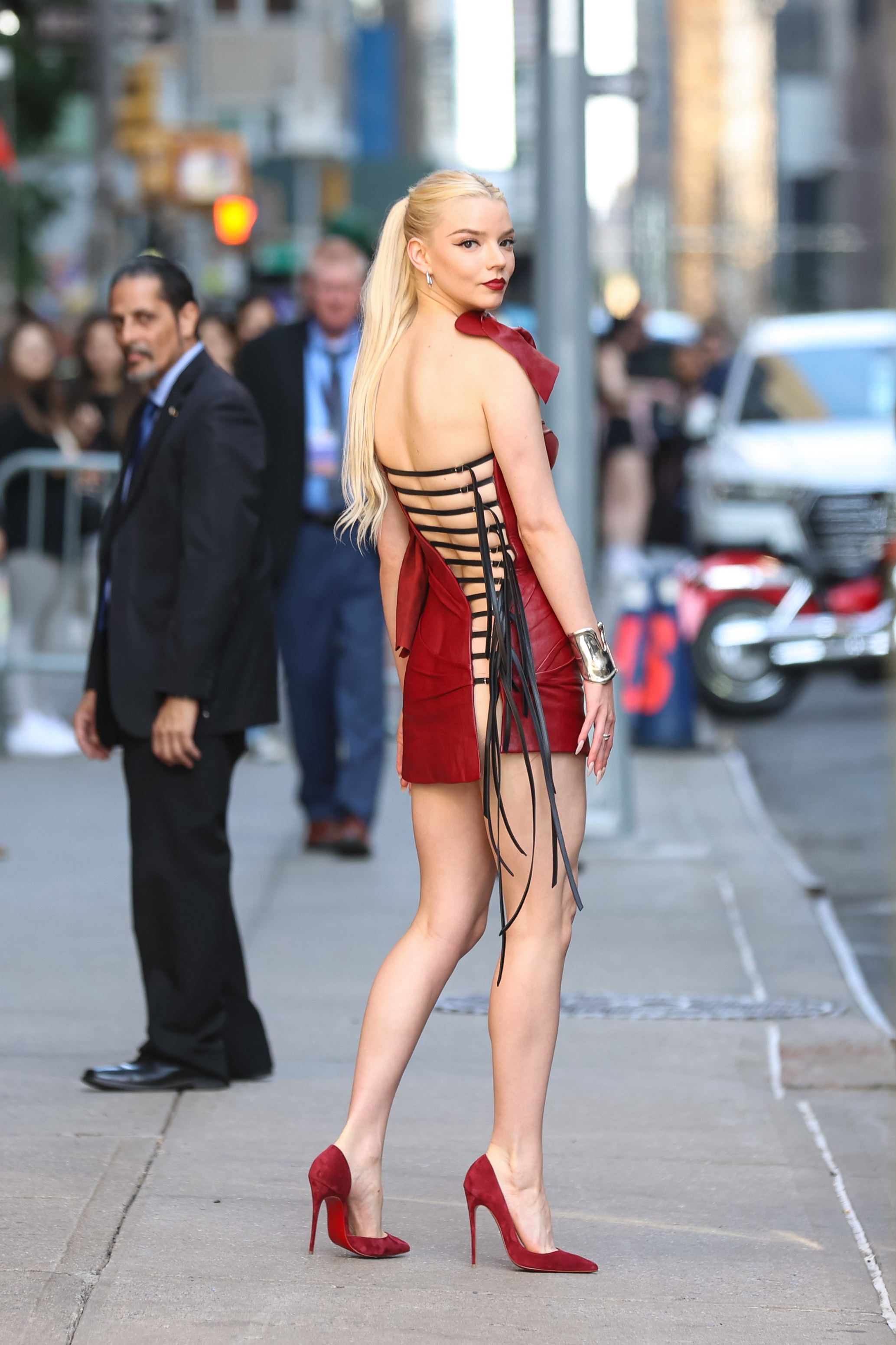 Anya Taylor-Joy on a city street wearing a backless red mini dress with black crisscross straps and red high heels, looking over her shoulder