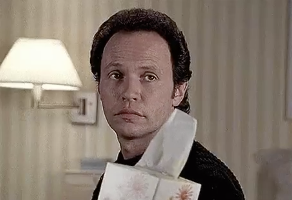 Billy Crystal holding a tissue box, looking off-screen with a solemn expression in a dimly lit room