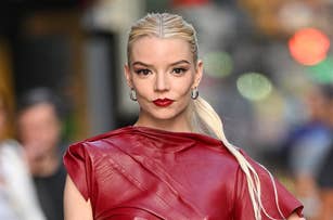 Anya Taylor-Joy poses for a photo wearing a stylish, sleeveless, textured red dress with a high neckline and unique wrap detail. Her hair is pulled back