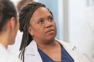 Chandra Wilson in a medical setting wearing a white lab coat and navy blue scrubs, looking attentively at someone off-camera