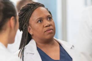 Chandra Wilson in a medical setting wearing a white lab coat and navy blue scrubs, looking attentively at someone off-camera