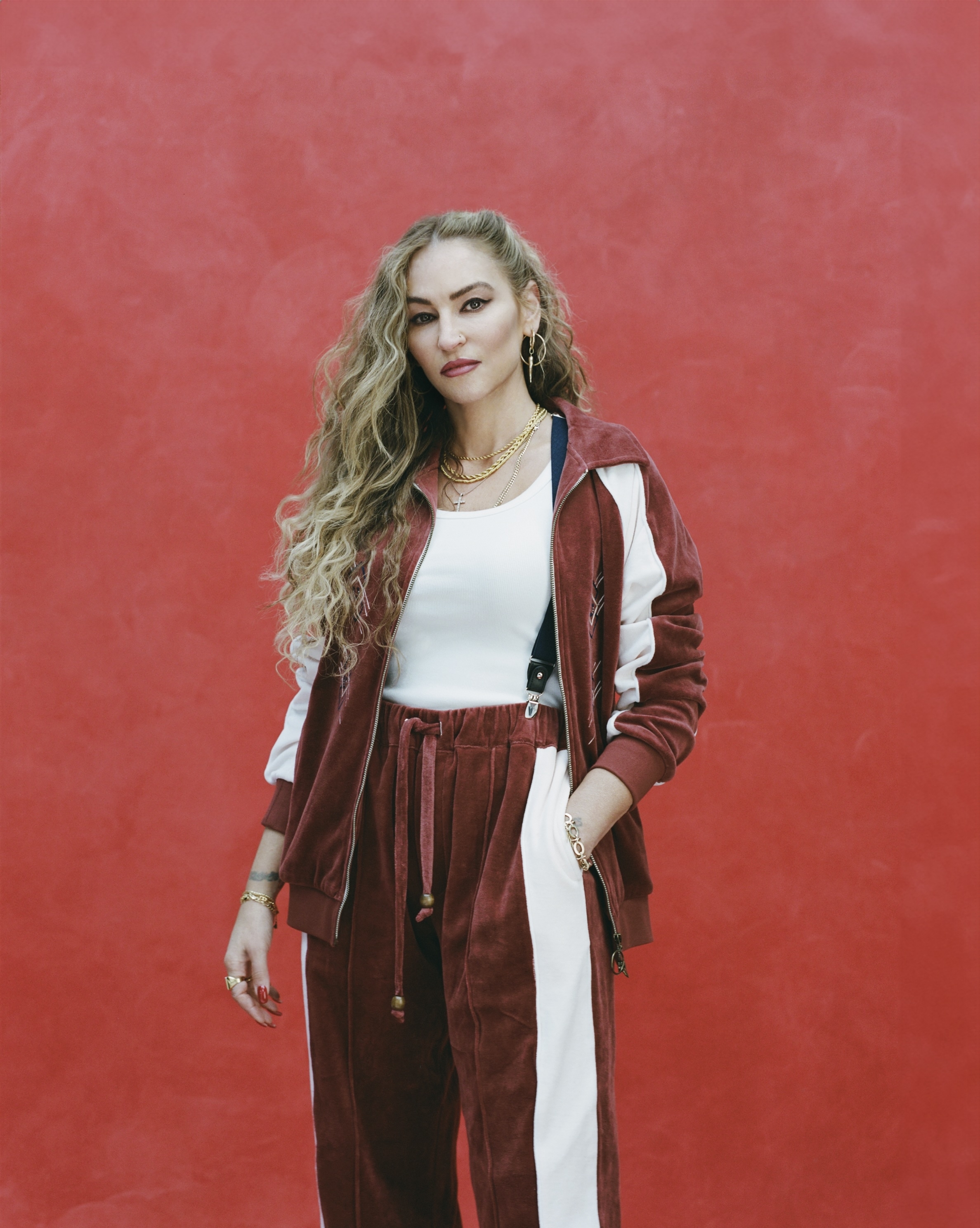 Woman with long curly hair wearing a velour tracksuit and a white tank top, standing with a confident expression against a red background