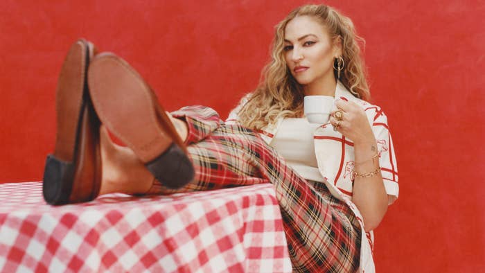 Woman poses casually, feet on a checkered table, wearing plaid pants and a printed shirt, holding a teacup