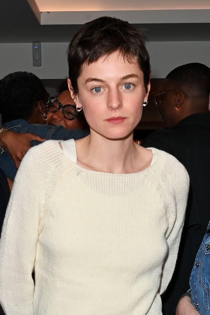 Emma Corrin wears a casual knit sweater at an event, standing in a crowd