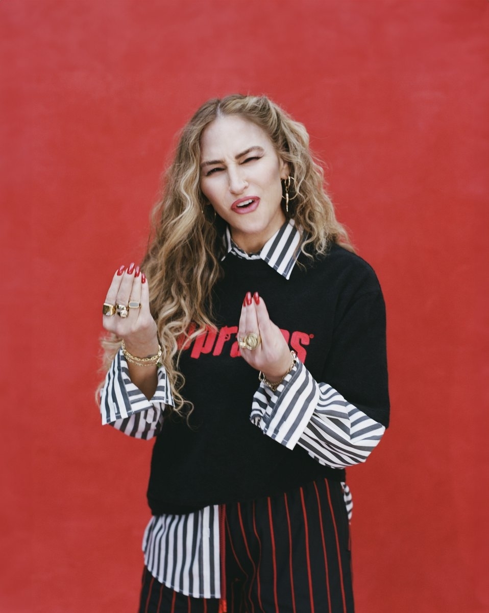 A woman stands in front of a red background, wearing a black sweater with stripes and &quot;popstar&quot; text, and striped pants. She has curly hair and red-painted nails, making expressive hand gestures