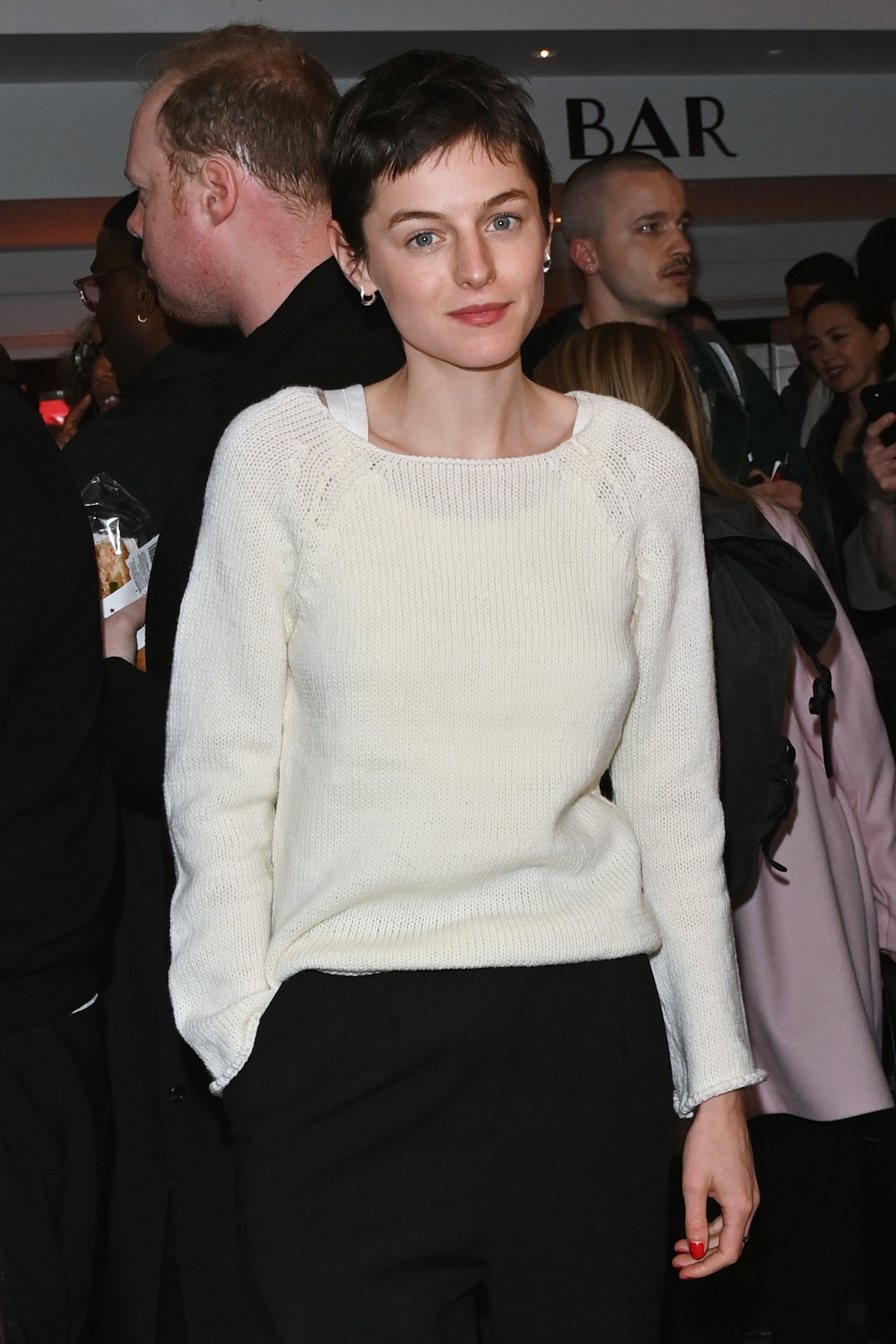 Emma Corrin in a white sweater with short hair, attended an event with a crowd in the background