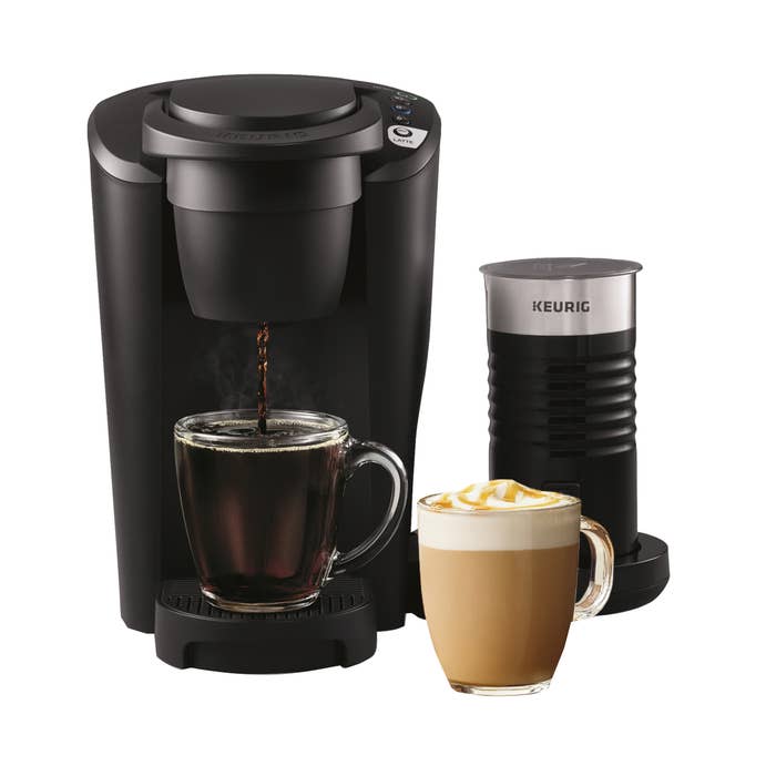 Keurig coffee maker brewing a cup of black coffee, with a milk frother beside it and a mug of frothy cappuccino