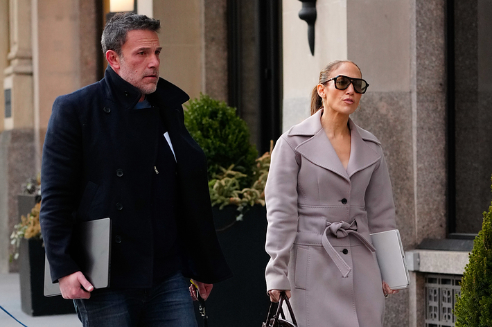 Ben Affleck in a coat and jeans walking with Jennifer Lopez in a trench coat and boots, both carrying laptops on a city street