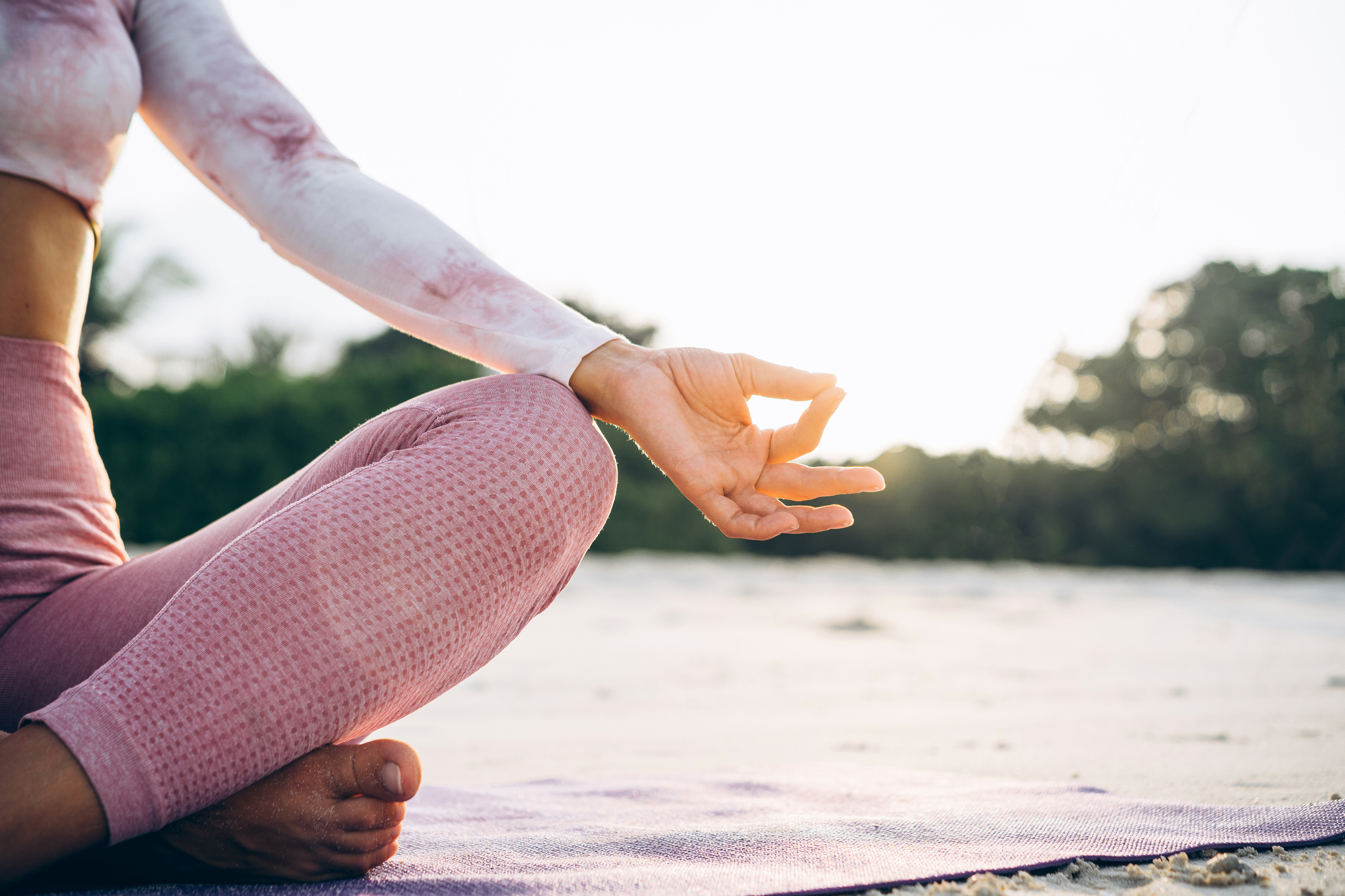 Person meditating outdoors on a beach while sitting cross-legged on a yoga mat, with hands in a mudra (gesture) position. Nearby, vegetation and a sunrise can be seen