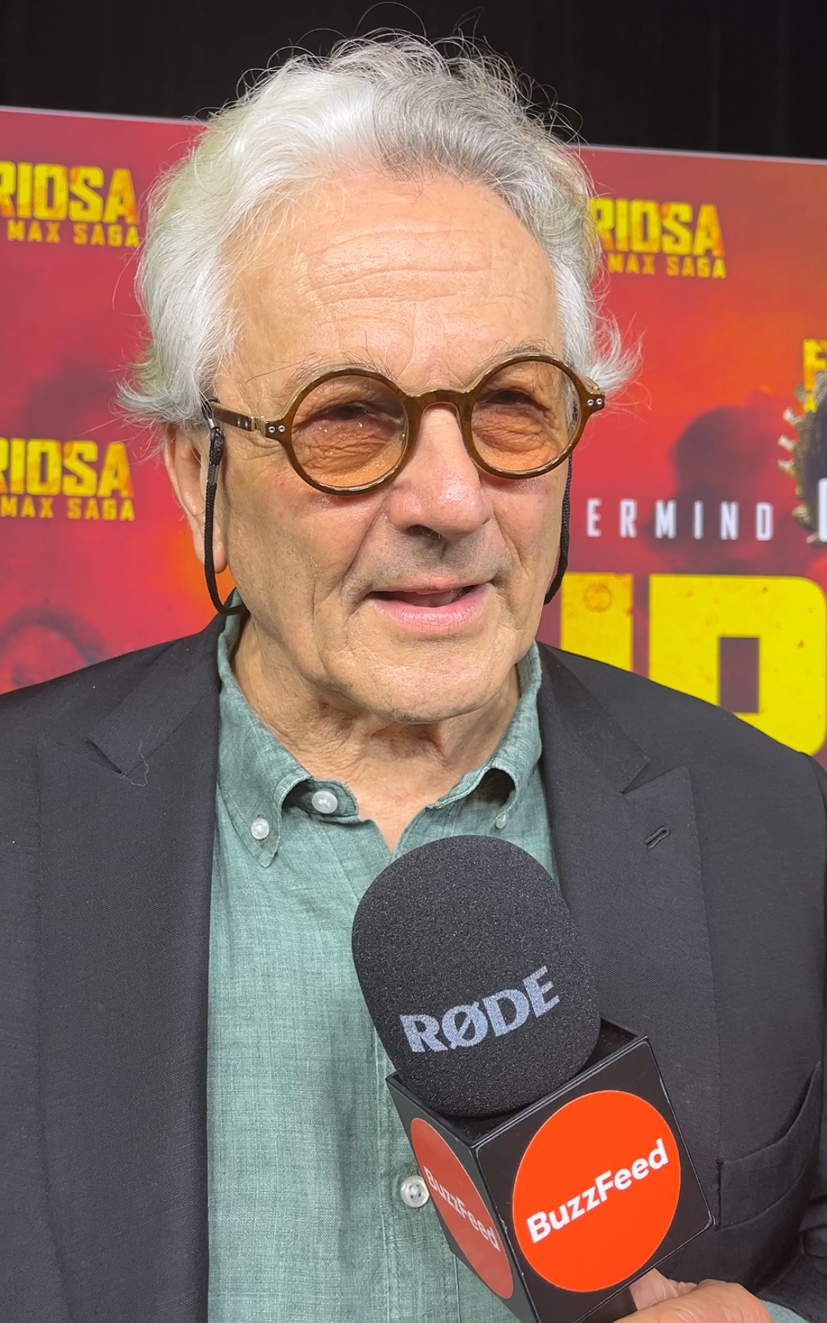 George Miller speaking to BuzzFeed with a RØDE microphone at "Furiosa" premiere. He's wearing round glasses and a casual shirt under a blazer