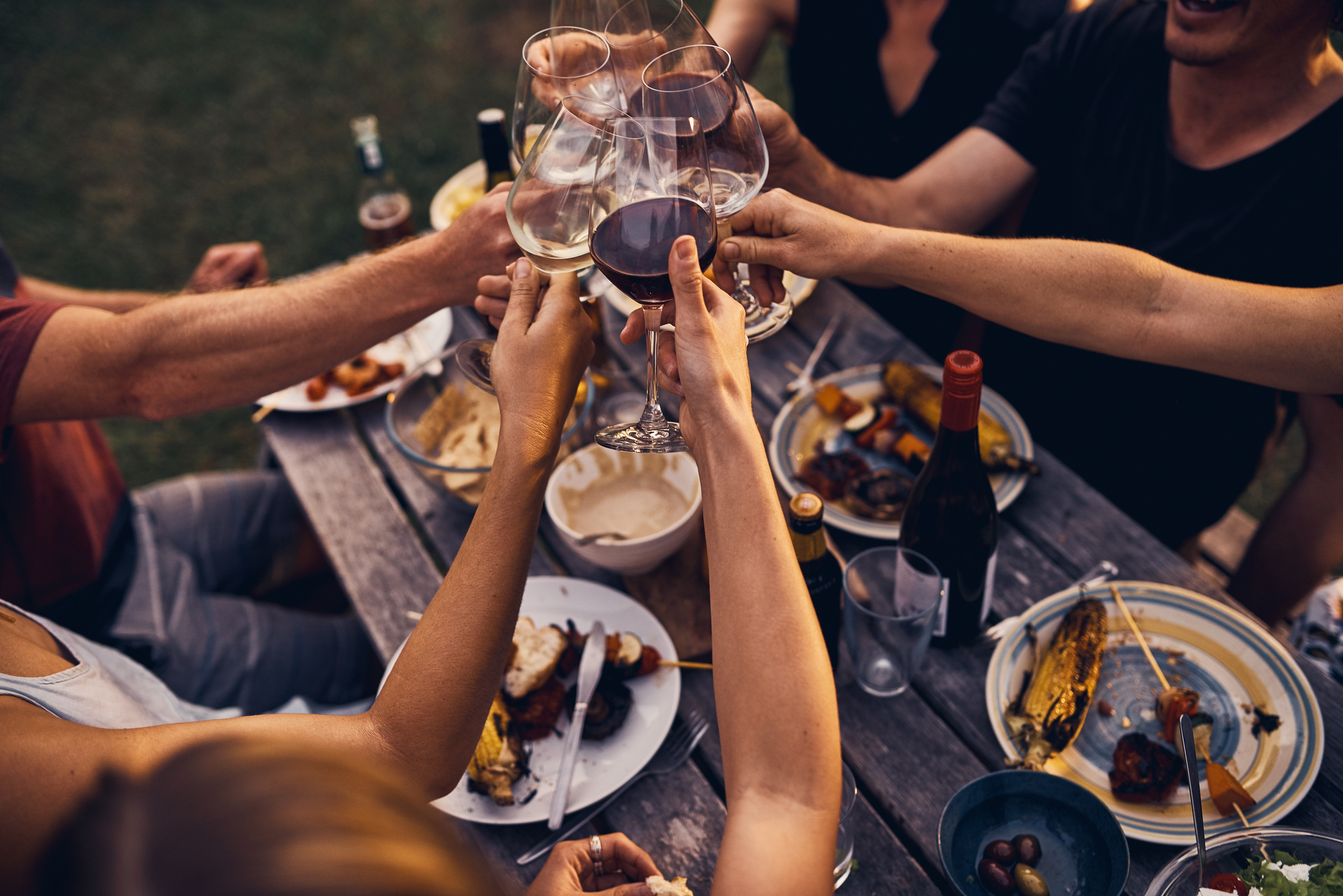 People raising wine glasses in a toast around a table with food and drinks. The image captures a festive, outdoor gathering. No identifiable individuals present