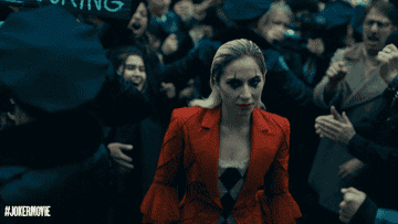 Lady Gaga, in a red suit with black eye makeup, walks through a crowd with police and fans behind fences, promoting #JokerMovie