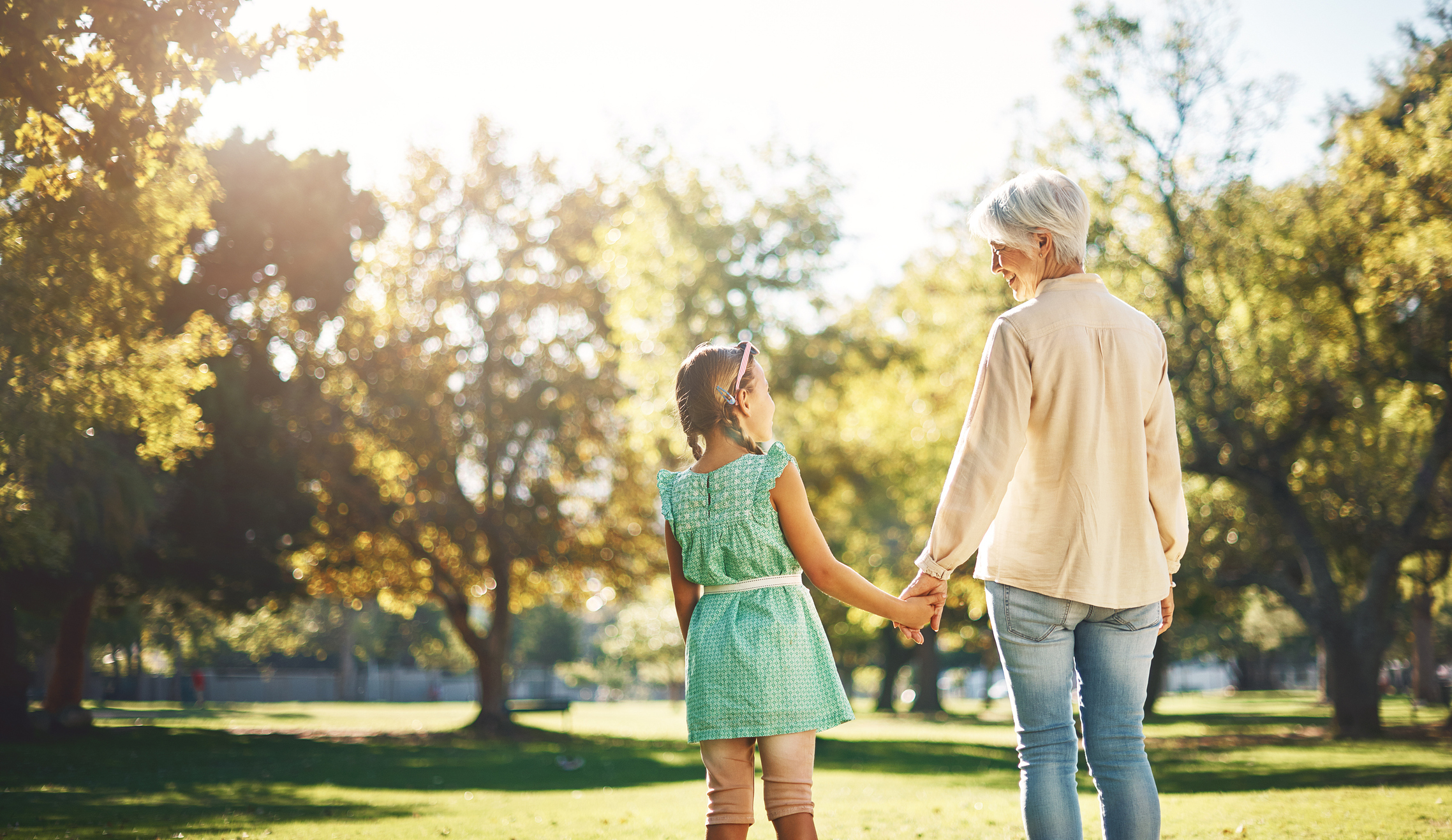 An older woman and a young girl walk hand-in-hand through a sunny park, smiling at each other. The woman wears jeans and a shirt, and the girl wears a dress and boots