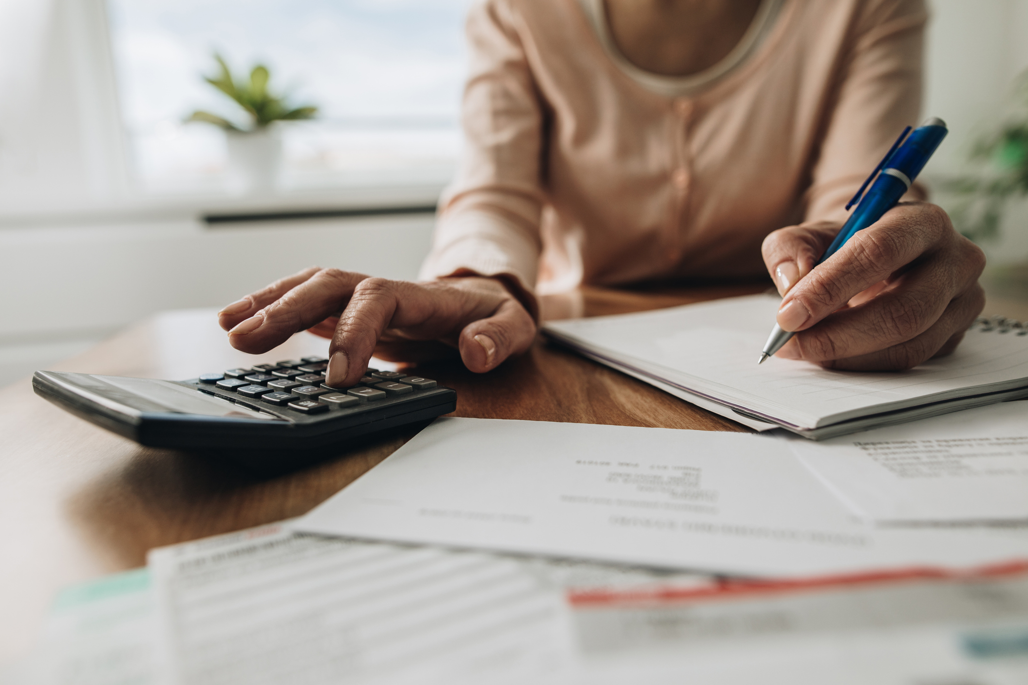 A person in a long-sleeve shirt uses a calculator with one hand and takes notes with the other, surrounded by documents on a desk