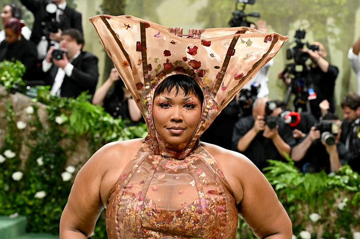 Lizzo poses on the red carpet in a floral-themed gown with a large headpiece, surrounded by photographers