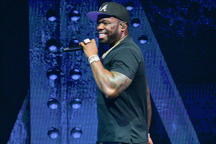 50 Cent smiling and performing on stage, wearing a black baseball cap and black t-shirt, holding a microphone