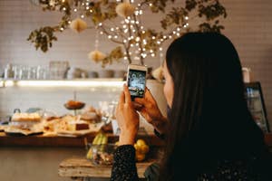 A person is holding a smartphone and taking a photo of a decorated table in a cozy café. Twinkling lights and hanging decorations create a warm atmosphere in the background