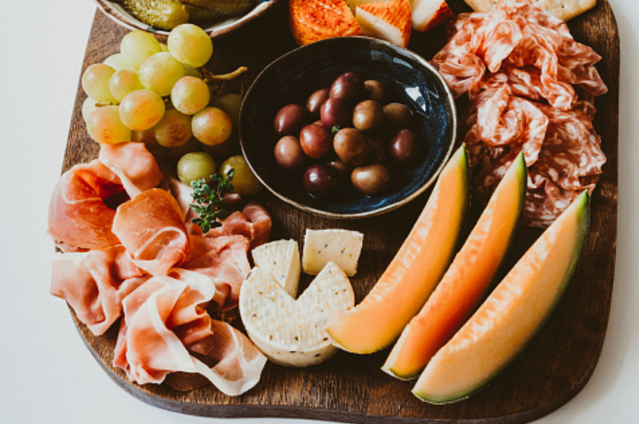 A charcuterie board featuring a variety of meats, cheeses, melons, grapes, olives, and pickles displayed on a wooden platter