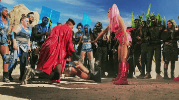 Group of people in a dramatic scene with costumed individuals. One person in red assists another on the ground, and a person in pink boots and outfit stands nearby