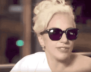 Lady Gaga wearing sunglasses and a white top, smiling in an indoor setting