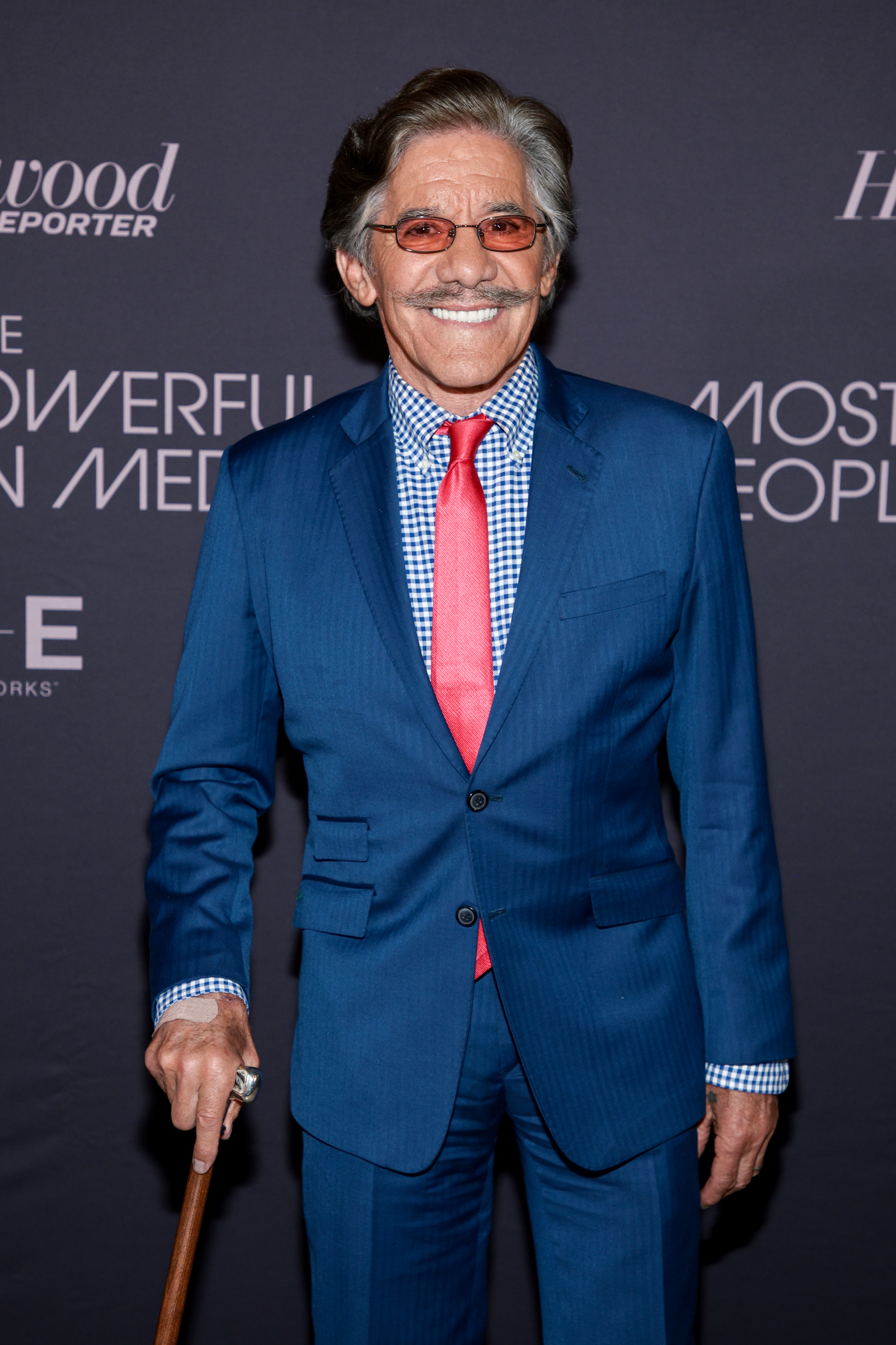 Geraldo Rivera smiles while wearing a blue suit with a red tie and a checkered shirt; he holds a cane and stands in front of a media event backdrop