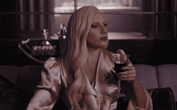 Lady Gaga in a satin gown, seated on a couch, holding a glass of wine, with a contemplative expression