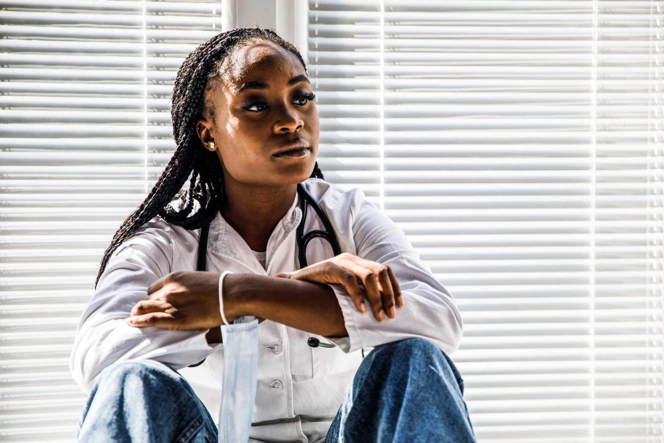 A woman with braided hair, wearing a white medical coat and a stethoscope, sits pensively in front of window blinds