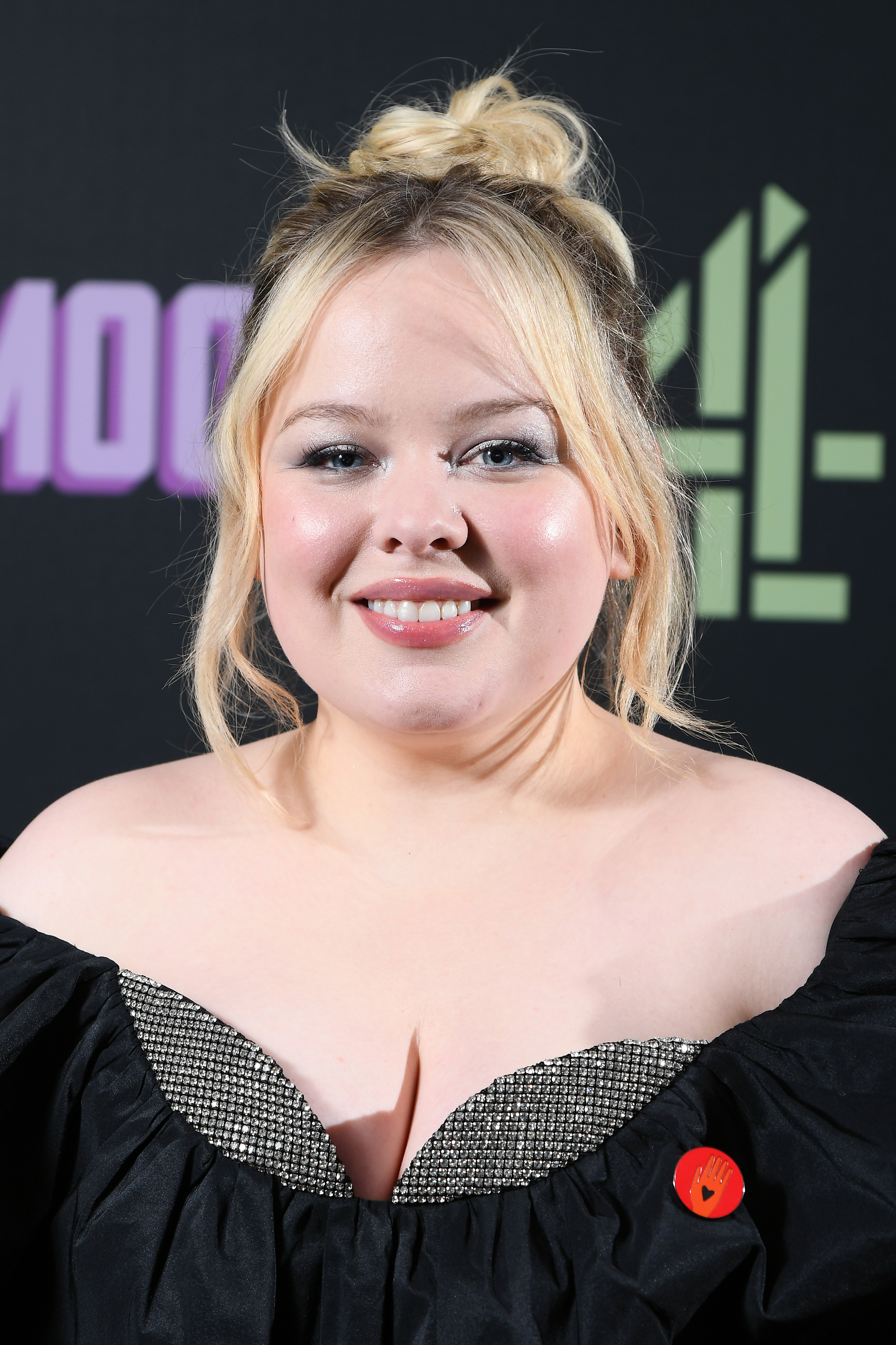 Nicola Coughlan smiles at the camera, wearing an off-shoulder dress with a patterned neckline and the pin