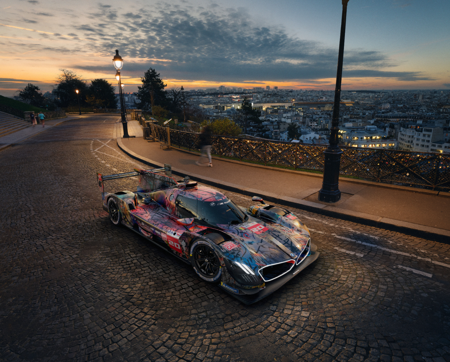 Race car on a cobblestone street in a city at sunset with city lights in the background. The car features a colorful, graffiti-style design