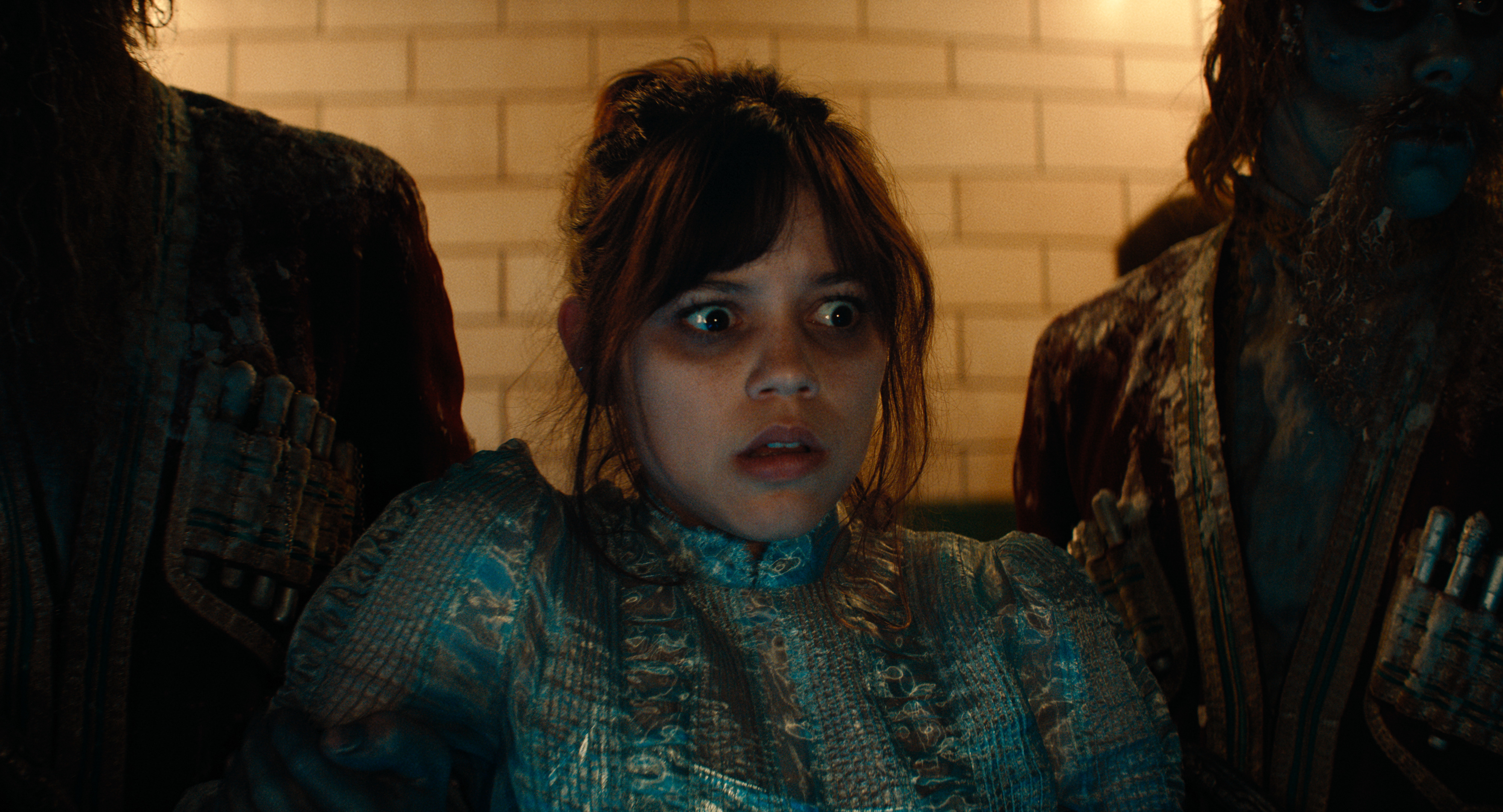 Jenna Ortega in an ornate, old-fashioned dress, looking startled while surrounded by two ghostly figures in a dimly lit setting