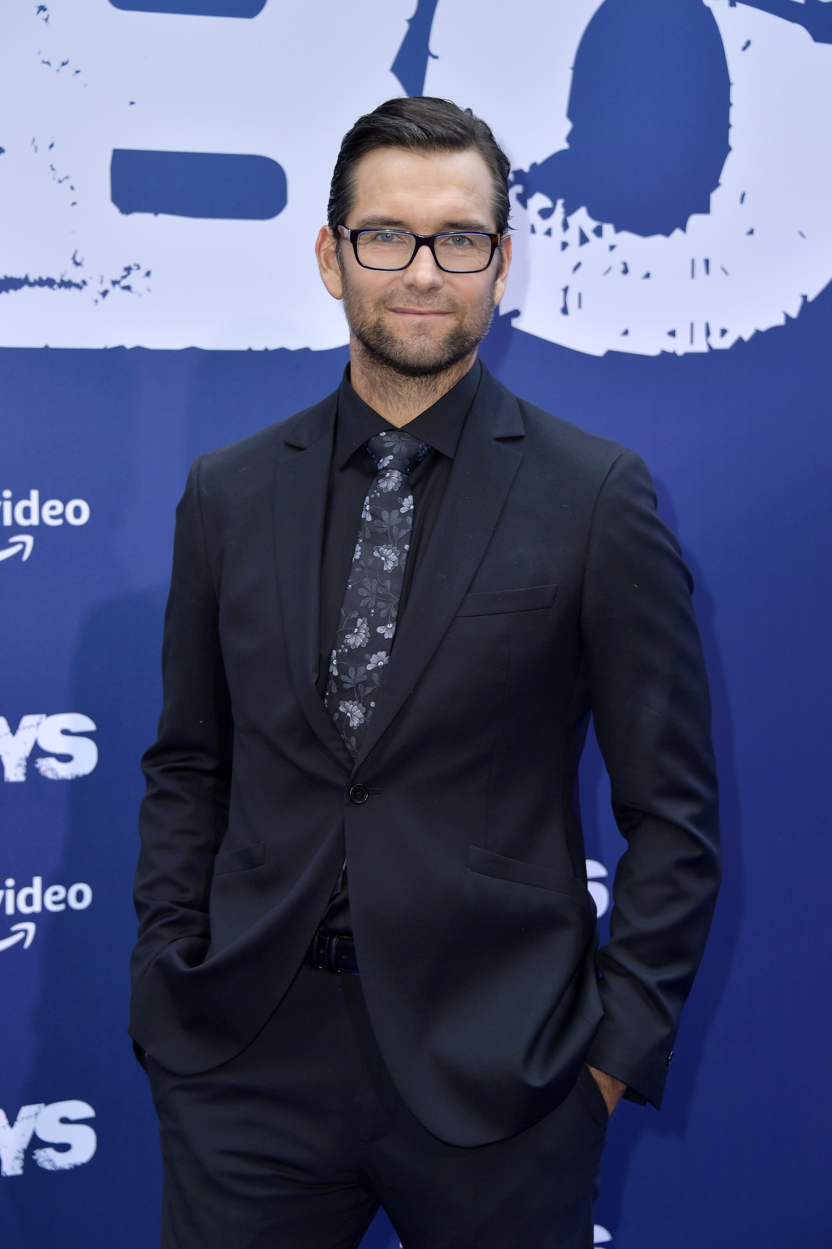 Man in dark suit with floral tie on a red carpet, hands in pockets, posing in front of a backdrop with text